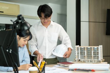 İki kadın ve erkek mühendis veya mimar çalışma masasında inşaat modeli ve ofis işyerindeki diğer ekipmanlarla birlikte çalışıyor. Erkek bir iş arkadaşı konuşurken inşaat projesinin planlarını, planlarını ve planlarını ele alıyor ve tartışıyorlar.