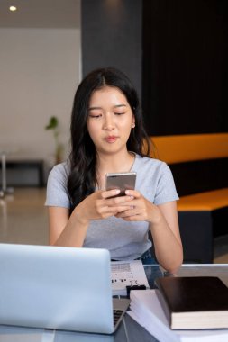 Genç bir kadın modern bir çalışma alanında oturuyor ve işine devam etmeden önce akıllı telefonuyla internete bağlanarak bilgisayarla ve kağıtlarla çalışmaya ara veriyor.. 