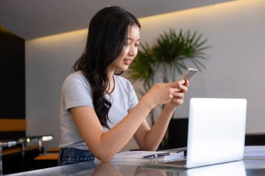 Genç bir kadın modern bir çalışma alanında oturuyor ve işine devam etmeden önce akıllı telefonuyla internete bağlanarak bilgisayarla ve kağıtlarla çalışmaya ara veriyor.. 