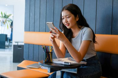 Genç bir kadın modern bir çalışma alanında oturuyor ve işine devam etmeden önce akıllı telefonuyla internete bağlanarak çalışmaya ya da çalışmaya ara veriyor.. 