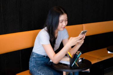 Genç bir kadın modern bir çalışma alanında oturuyor ve işine devam etmeden önce akıllı telefonuyla internete bağlanarak çalışmaya ya da çalışmaya ara veriyor.. 