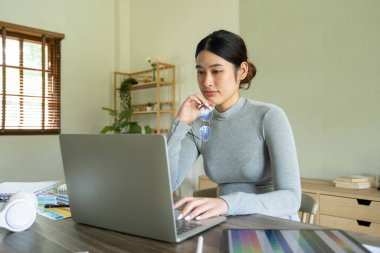 Genç bayan mimar ya da iç mimar ofisinde ekipmanlarla masasında oturuyor, dizüstü bilgisayarla çalışıyor ve tasarım fikirlerini hayata geçiriyor ve çizelgelerden renkler seçiyor, tüm projeyi hayal ederek. 