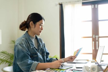 Genç bayan mimar veya iç mimar, elinde tabletle ofisinde masasında oturuyor, dizüstü bilgisayardaki tasarım fikirlerini çiziyor ve hayata geçiriyor ve çizelgelerden renkler seçiyor, tüm projeyi öngörüyor.. 