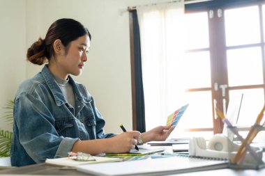Genç bayan mimar veya iç mimar, elinde tabletle ofisinde masasında oturuyor, dizüstü bilgisayardaki tasarım fikirlerini çiziyor ve hayata geçiriyor ve çizelgelerden renkler seçiyor, tüm projeyi öngörüyor.. 
