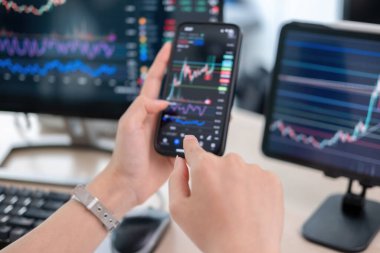 Genç bir kadın, bilgisayar, dizüstü bilgisayar ve tabletle çevrili çalışma masasında özenle çalışıyor. Akıllı telefonunun ekranında gösterilen hisse senedi grafiklerini inceliyor, potansiyel yatırım fırsatları için verileri analiz ediyor..