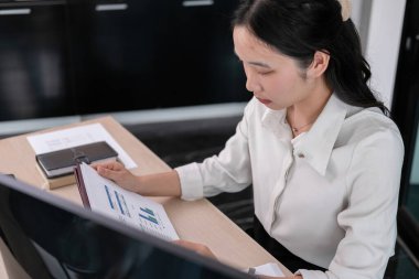 Etrafı bilgisayar, dizüstü bilgisayar ve iPad ile çevrili genç bir kadın ofis masasında özenle çalışıyor. Ekranda gösterilen hisse senedi grafiklerini dikkatle inceler ve potansiyel yatırım fırsatları için verileri analiz eder..