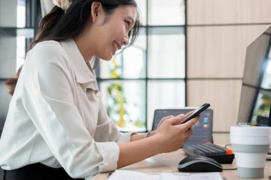 Genç bir kadın, bir bilgisayar ve tabletle çevrili ofis masasında özenle çalışıyor. Ekranda gösterilen hisse senedi grafiklerini dikkatle inceler ve akıllı telefon kullanır, potansiyel yatırım fırsatları için verileri analiz eder..
