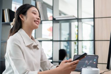 Genç bir kadın, bir bilgisayar ve tabletle çevrili ofis masasında özenle çalışıyor. Ekranda gösterilen hisse senedi grafiklerini dikkatle inceler ve akıllı telefon kullanır, potansiyel yatırım fırsatları için verileri analiz eder..