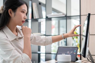 Etrafı bilgisayar ve iPad ile çevrili genç bir kadın ofis masasında özenle çalışıyor. Ekranda gösterilen hisse senedi grafiklerini dikkatle inceler ve inceler, potansiyel yatırım fırsatları için verileri analiz eder..