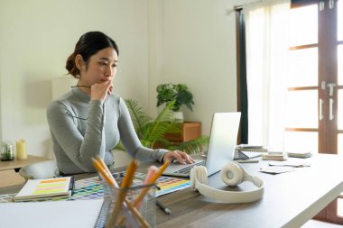 Genç bayan mimar ya da iç mimar ofisinde ekipmanlarla masasında oturuyor, dizüstü bilgisayarla çalışıyor ve tasarım fikirlerini hayata geçiriyor ve çizelgelerden renkler seçiyor, tüm projeyi hayal ederek. 