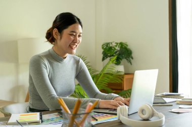 Genç bayan mimar ya da iç mimar ofisinde ekipmanlarla masasında oturuyor, dizüstü bilgisayarla çalışıyor ve tasarım fikirlerini hayata geçiriyor ve tüm projeyi öngören çizelgelerden renkleri seçiyor.. 