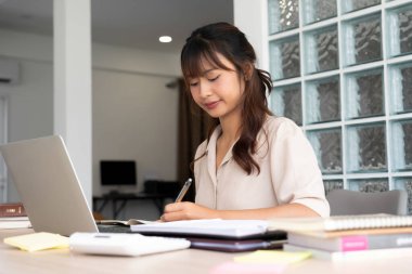 Genç bir kadın işyerinde özenle oturuyor, çevrimiçi çalışmaya dalmış ve güvenilir dizüstü bilgisayarıyla bir okul projesi için araştırma yapıyor ve notlar yazıyor..