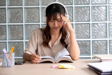 Genç bir kadın ya da öğrenci, elinde notlar ve kitaplarla çalışma alanında oturmuş, kulaklıkla müzik eşliğinde verimlilik ve rahatlama üzerine bir kitap okumaktan keyif alıyor..