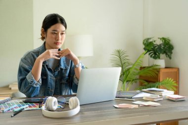 Genç bayan mimar ya da iç mimar ofisinde ekipmanlarla masasında oturuyor, dizüstü bilgisayarla çalışıyor ve tasarım fikirlerini hayata geçiriyor ve çizelgelerden renkler seçiyor, tüm projeyi hayal ederek. 
