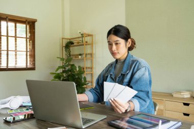 Genç bayan mimar veya iç mimar, elinde tabletle ofisinde masasında oturuyor, dizüstü bilgisayardaki tasarım fikirlerini çiziyor ve hayata geçiriyor ve çizelgelerden renkler seçiyor, tüm projeyi öngörüyor.. 