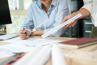 İki erkek ve kadın mühendis veya mimar çalışma masasında kağıt işleri ve ofis işyerindeki diğer ekipmanlarla birlikte çalışarak bir cetveli işaret ediyor, inşaat projesinin planları hakkında plan yapıyor ve tartışıyorlar..