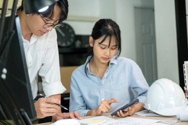 İki erkek ve kadın mühendis ya da mimar çalışma masasında profesyonel ekipmanlarla birlikte çalışarak inşaat projesinin planlarını gösteriyorlar, çiziyorlar, planlıyorlar ve tartışıyorlar..