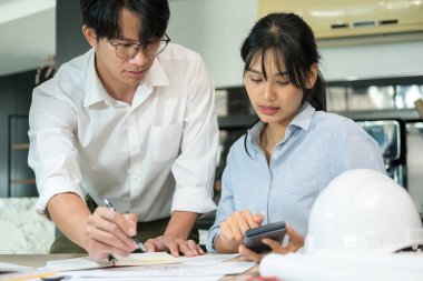 İki erkek ve kadın mühendis, müteahhit veya mimar birlikte çalışıyor, inşaat projesinin grafik tasarımını çiziyor ve kağıt ve hesap makinesiyle birlikte çalışma masasında gelişim için tartışıyorlar. Mühendislik ve Takım Çalışması konsepti.