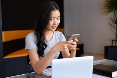 Genç bir kadın modern bir çalışma alanında oturuyor ve işine devam etmeden önce akıllı telefonuyla internete bağlanarak bilgisayarla ve kağıtlarla çalışmaya ara veriyor.. 