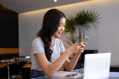 Genç bir kadın modern bir çalışma alanında oturuyor ve işine devam etmeden önce akıllı telefonuyla internete bağlanarak bilgisayarla ve kağıtlarla çalışmaya ara veriyor.. 