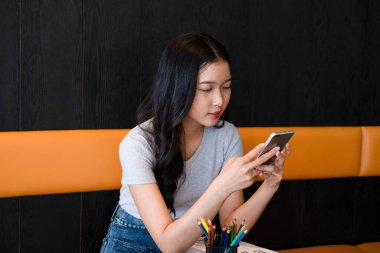 Genç bir kadın modern bir çalışma alanında oturuyor ve işine devam etmeden önce akıllı telefonuyla internete bağlanarak çalışmaya ya da çalışmaya ara veriyor.. 