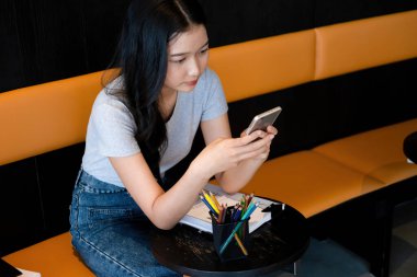 Genç bir kadın modern bir çalışma alanında oturuyor ve işine devam etmeden önce akıllı telefonuyla internete bağlanarak çalışmaya ya da çalışmaya ara veriyor.. 