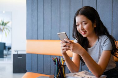 Genç bir kadın modern bir çalışma alanında oturuyor ve işine devam etmeden önce akıllı telefonuyla internete bağlanarak çalışmaya ya da çalışmaya ara veriyor.. 