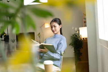 Genç Asyalı kadın pencerenin yanında kitap okuyarak huzurlu bir anın tadını çıkarıyor. Sakin yaşam tarzı, kendi kendine öğrenme ve evdeki farkındalık..