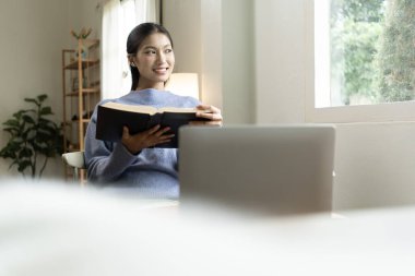 Genç Asyalı kadın pencerenin yanında kitap okuyarak huzurlu bir anın tadını çıkarıyor. Sakin yaşam tarzı, kendi kendine öğrenme ve evdeki farkındalık..