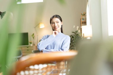 Genç Asyalı kadın pencerenin yanında kitap okuyarak huzurlu bir anın tadını çıkarıyor. Sakin yaşam tarzı, kendi kendine öğrenme ve evdeki farkındalık..