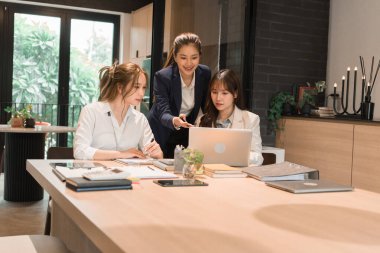 Üç genç iş kadınından oluşan bir grup, belgeler, tablet, dizüstü bilgisayar ve diğer ekipmanlarla birlikte bir masada oturuyorlar. Şirketi geliştirmek için bir tartışma ve planlama oturumu yapıyorlar..