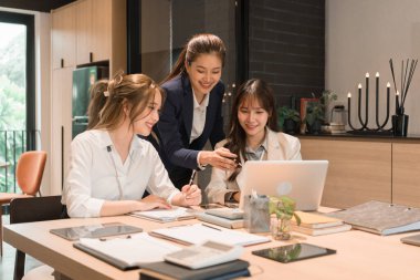 Üç genç iş kadınından oluşan bir grup, belgeler, tablet, dizüstü bilgisayar ve diğer ekipmanlarla birlikte bir masada oturuyorlar. Şirketi geliştirmek için bir tartışma ve planlama oturumu yapıyorlar..