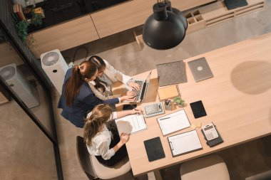 Üç genç iş kadınından oluşan bir grup, belgeler, tablet, dizüstü bilgisayar ve diğer ekipmanlarla birlikte bir masada oturuyorlar. Şirketi geliştirmek için bir tartışma ve planlama oturumu yapıyorlar..