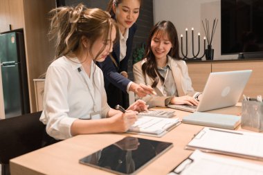 Üç genç iş kadınından oluşan bir grup, belgeler, tablet, dizüstü bilgisayar ve diğer ekipmanlarla birlikte bir masada oturuyorlar. Şirketi geliştirmek için bir tartışma ve planlama oturumu yapıyorlar..