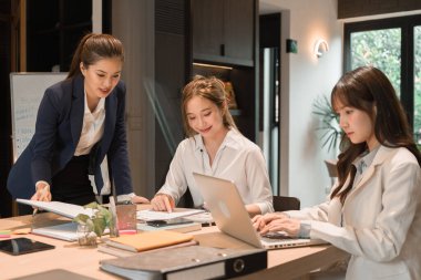 Üç genç iş kadınından oluşan bir grup, belgeler, tablet, dizüstü bilgisayar ve diğer ekipmanlarla birlikte bir masada oturuyorlar. Şirketi geliştirmek için bir tartışma ve planlama oturumu yapıyorlar..