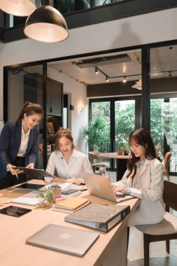 Üç genç iş kadınından oluşan bir grup, belgeler, tablet, dizüstü bilgisayar ve diğer ekipmanlarla birlikte bir masada oturuyorlar. Şirketi geliştirmek için bir tartışma ve planlama oturumu yapıyorlar..