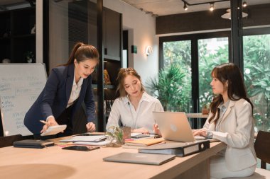 Üç genç iş kadınından oluşan bir grup, belgeler, tablet, dizüstü bilgisayar ve diğer ekipmanlarla birlikte bir masada oturuyorlar. Şirketi geliştirmek için bir tartışma ve planlama oturumu yapıyorlar..