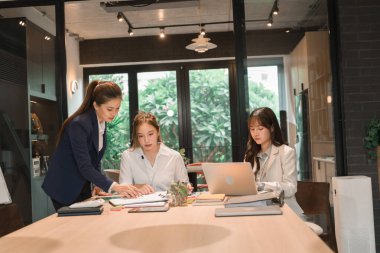 Üç genç iş kadınından oluşan bir grup, belgeler, tablet, dizüstü bilgisayar ve diğer ekipmanlarla birlikte bir masada oturuyorlar. Şirketi geliştirmek için bir tartışma ve planlama oturumu yapıyorlar..