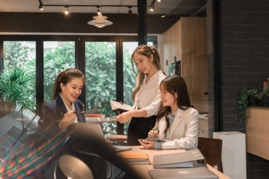 Üç genç iş kadınından oluşan bir grup, belgeler, tablet, dizüstü bilgisayar ve diğer ekipmanlarla birlikte bir masada oturuyorlar. Şirketi geliştirmek için bir tartışma ve planlama oturumu yapıyorlar..