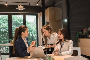 Üç genç iş kadınından oluşan bir grup, belgeler, tablet, dizüstü bilgisayar ve diğer ekipmanlarla birlikte bir masada oturuyorlar. Şirketi geliştirmek için bir tartışma ve planlama oturumu yapıyorlar..