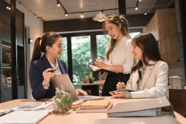 Üç genç iş kadınından oluşan bir grup, belgeler, tablet, dizüstü bilgisayar ve diğer ekipmanlarla birlikte bir masada oturuyorlar. Şirketi geliştirmek için bir tartışma ve planlama oturumu yapıyorlar..
