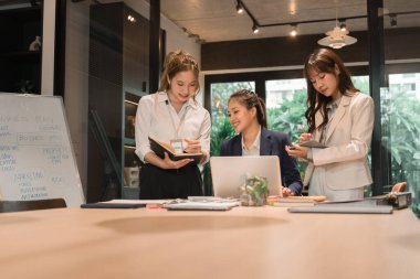 Üç genç iş kadınından oluşan bir grup, belgeler, tablet, dizüstü bilgisayar ve diğer ekipmanlarla birlikte bir masada oturuyorlar. Şirketi geliştirmek için bir tartışma ve planlama oturumu yapıyorlar..