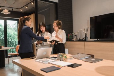 Üç genç iş kadınından oluşan bir grup, belgeler, tablet, dizüstü bilgisayar ve diğer ekipmanlarla birlikte bir masada oturuyorlar. Şirketi geliştirmek için bir tartışma ve planlama oturumu yapıyorlar..