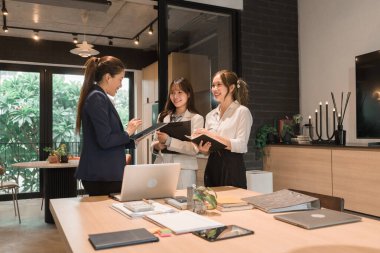 Üç genç iş kadınından oluşan bir grup, belgeler, tablet, dizüstü bilgisayar ve diğer ekipmanlarla birlikte bir masada oturuyorlar. Şirketi geliştirmek için bir tartışma ve planlama oturumu yapıyorlar..