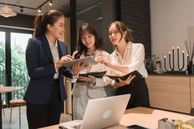 Üç genç iş kadınından oluşan bir grup, belgeler, tablet, dizüstü bilgisayar ve diğer ekipmanlarla birlikte bir masada oturuyorlar. Şirketi geliştirmek için bir tartışma ve planlama oturumu yapıyorlar..