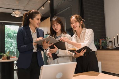 Üç genç iş kadınından oluşan bir grup, belgeler, tablet, dizüstü bilgisayar ve diğer ekipmanlarla birlikte bir masada oturuyorlar. Şirketi geliştirmek için bir tartışma ve planlama oturumu yapıyorlar..
