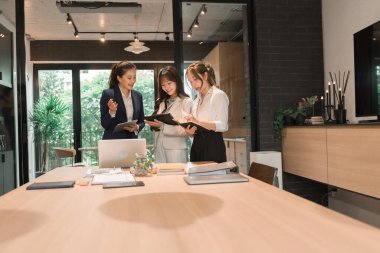 Üç genç iş kadınından oluşan bir grup, belgeler, tablet, dizüstü bilgisayar ve diğer ekipmanlarla birlikte bir masada oturuyorlar. Şirketi geliştirmek için bir tartışma ve planlama oturumu yapıyorlar..