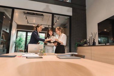 Üç genç iş kadınından oluşan bir grup, belgeler, tablet, dizüstü bilgisayar ve diğer ekipmanlarla birlikte bir masada oturuyorlar. Şirketi geliştirmek için bir tartışma ve planlama oturumu yapıyorlar..