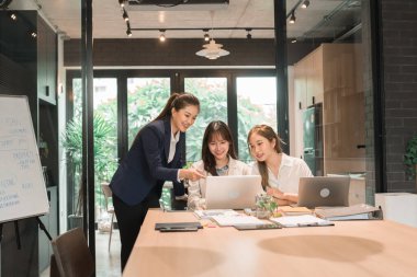 Üç genç iş kadınından oluşan bir grup, belgeler, tablet, dizüstü bilgisayar ve diğer ekipmanlarla birlikte bir masada oturuyorlar. Şirketi geliştirmek için bir tartışma ve planlama oturumu yapıyorlar..