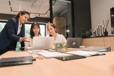 Üç genç iş kadınından oluşan bir grup, belgeler, tablet, dizüstü bilgisayar ve diğer ekipmanlarla birlikte bir masada oturuyorlar. Şirketi geliştirmek için bir tartışma ve planlama oturumu yapıyorlar..
